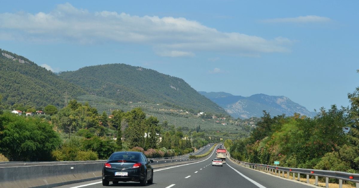 Imaginea prezintă un autoturism negru care circulă pe o autostradă ce șerpuiește printr-un peisaj cu dealuri împădurite, sub un cer senin de vară.