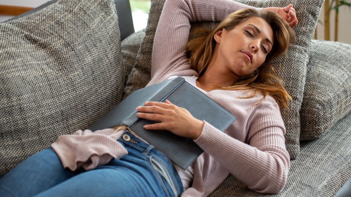 O tânără doarme pe canapea după-amiază, cu o carte deschisă pe abdomen