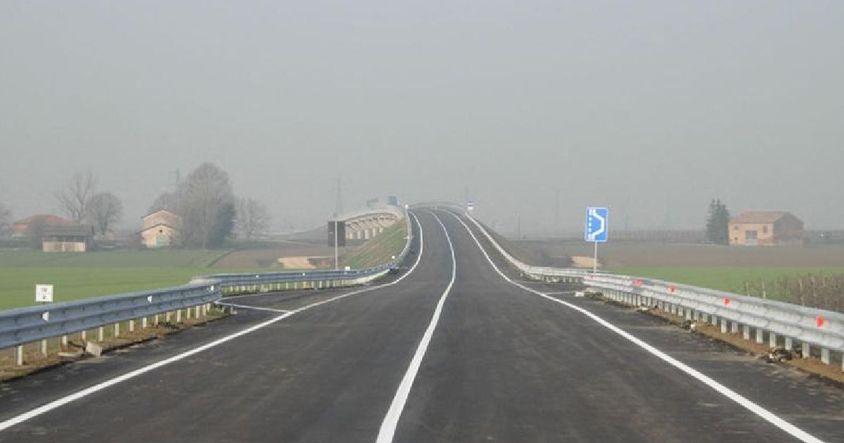 Un șosea cu ceață, goală, cu marcaje albe pe carosabil, flancată de parapete metalice și ferme și copaci îndepărtați de o parte și de alta.