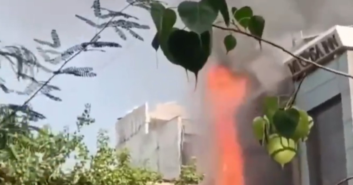 În imagine se observă un incendiu puternic izbucnit în apropierea unei clădiri, cu flăcări înalte și un nor dens de fum care se ridică deasupra zonei. Vegetația din prim-plan contrastează cu scena dramatică, surprinsă în timpul intervenției sau la scurt timp după izbucnirea focului.