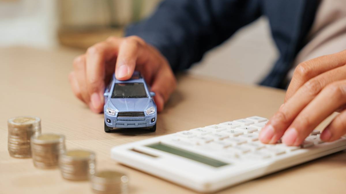 O persoană ține o machetă de mașină albastră într-o mână, lângă este un calculator de birou. Pe masă sunt așezate grămezi de monede, sugerând calcule financiare