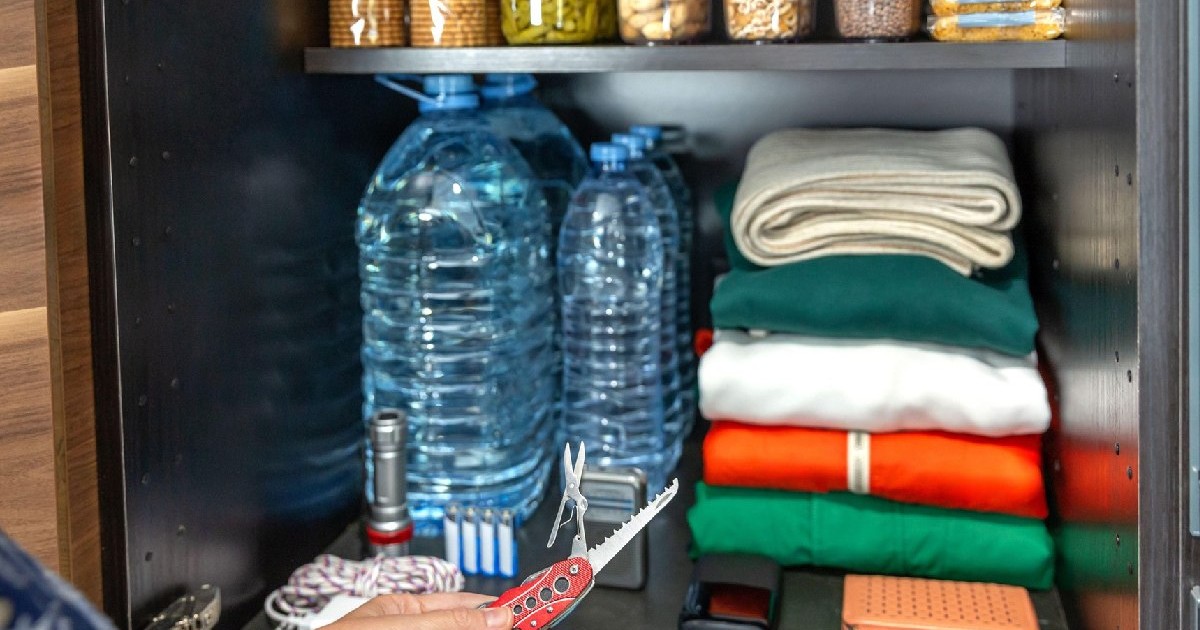 Mână de femeie ținând un cuțit multifuncțional lângă dulapul de urgență cu apă, haine, lanternă, baterii și powerbank. Femeia a pregătit un kit de supraviețuire pentru 72 de ore în caz de dezastre sau război.