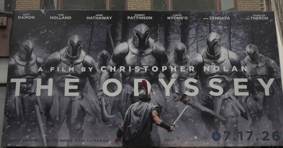 Imaginea surprinde un panou publicitar de mari dimensiuni pentru filmul „The Odyssey”, regizat de Christopher Nolan, amplasat pe fațada unei clădiri. Afișul prezintă un războinic în fața unor soldați în armură.