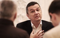 sorin grindeanu, cu mana pe piept și capul ușor într-o parte, purtând un costum negru cu cămașă albă, văzut printre umerii a alți doi bărbați îmbrăcați în costum