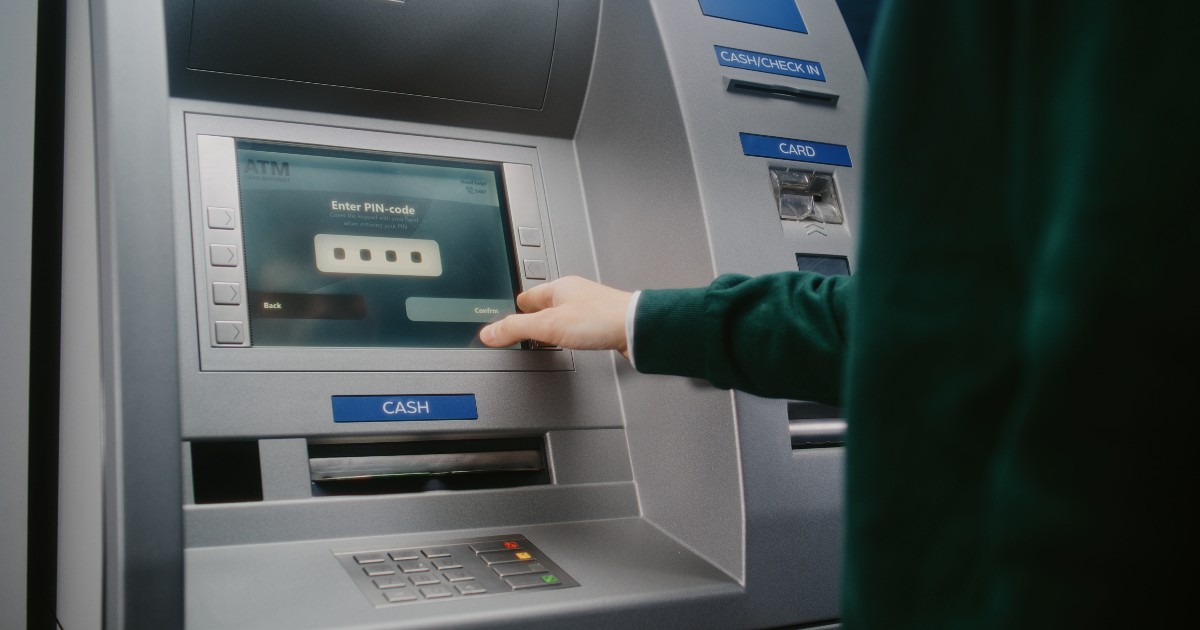 Un bărbat care scoate bani din bancomat cu cardul, poartă un pulover verde și introduce codul PIN