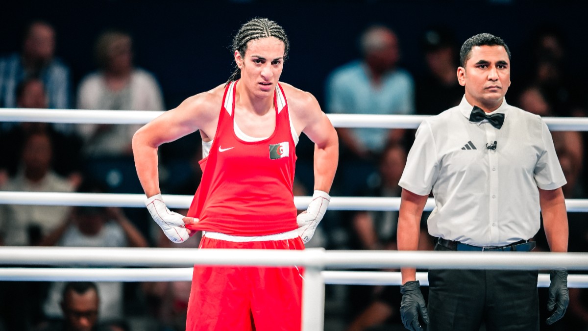 În imagine este o persoană îmbrăcată în haine roșii, într-un ring de box. Lângă aceasta se află un bărbat în cămașă albă.