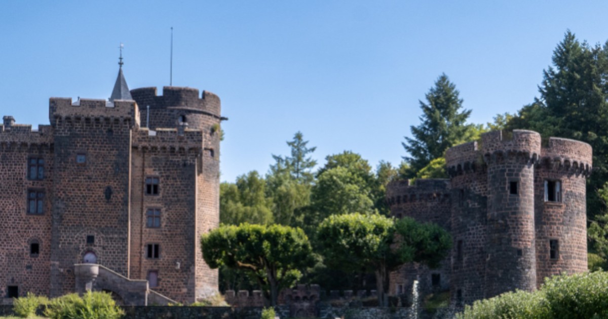 Imaginea prezintă un castel vechi din piatră, cu turnuri rotunde și ziduri groase, specific arhitecturii medievale. În jurul castelului se văd copaci și vegetație, iar în față se află o alee care duce spre intrare.