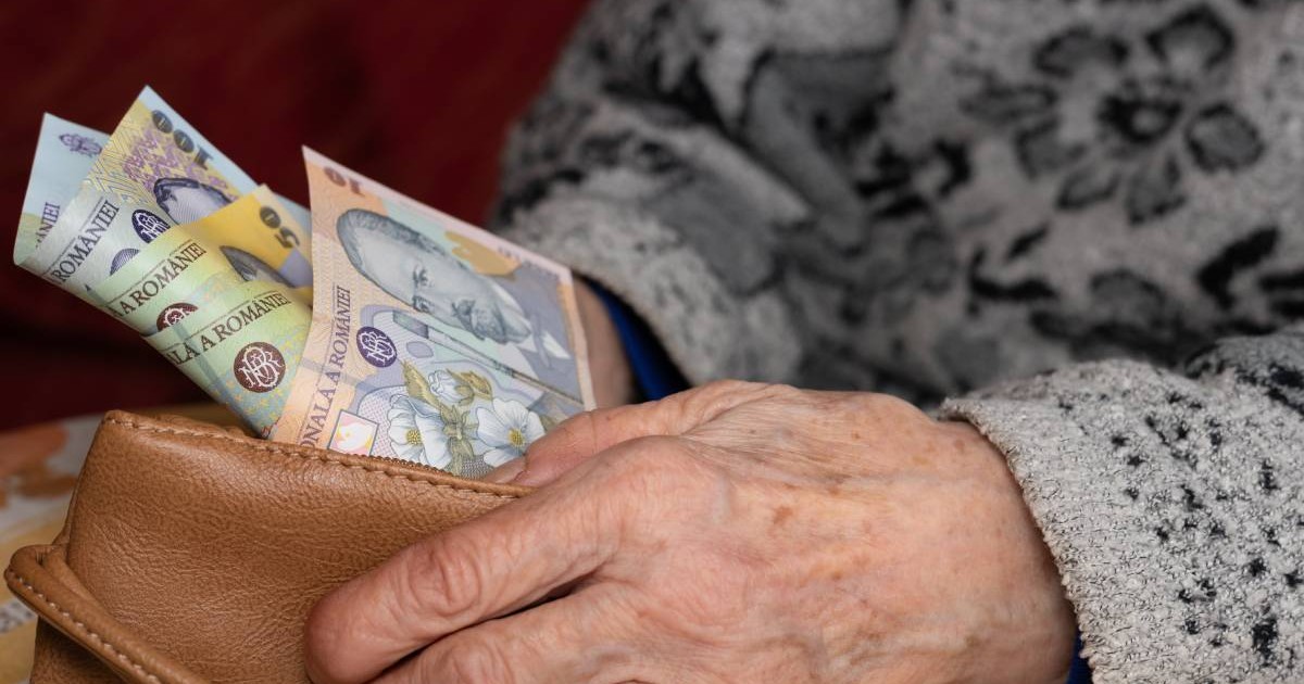O femeie în vârstă care stă jos și își numără banii din portofel. Femeia poartă un pulover gri cu un model, portofelul pe care îl ține în mână este din piele maro, iar din portofel ies bancnote de 100, 50 și 10 lei.