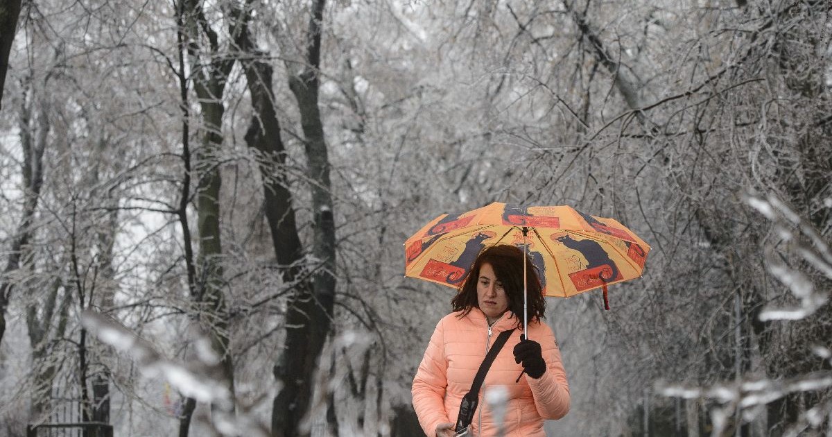 O femeie cu părul închis la culoare ține o umbrelă colorată, purtând o jachetă portocalie și mănuși negre, în timp ce merge pe o alee, într-un parc.
