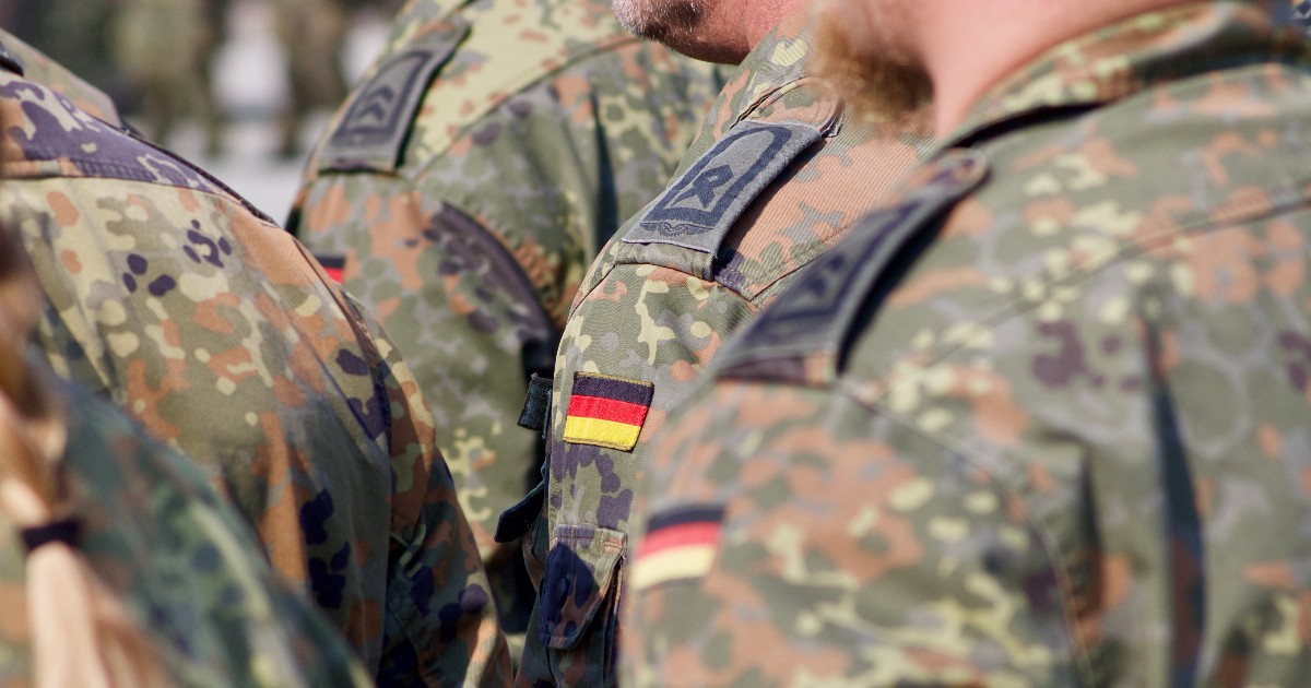Această imagine arată un grup de soldați (membri ai forțelor armate) îmbrăcați în uniforme de camuflaj, surprinși din spate. Pe umărul unuia dintre ei este vizibil un plasture (patch) cu culorile steagului Germaniei (negru, roșu și auriu), indicând faptul că sunt militari germani (Bundeswehr).