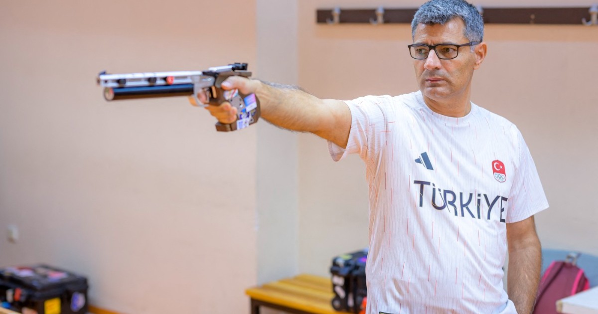 Imaginea îl surprinde pe Yusuf Dikeç, un sportiv turc, concentrat în timpul unui antrenament de tir, țintind cu un pistol sportiv cu aer comprimat. El poartă un tricou alb cu inscripția TÜRKİYE (Turcia) și simbolurile olimpice, într-un poligon de tragere interior.