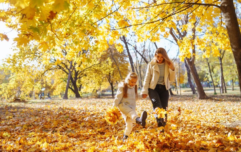 O mamă și fiica ei se joacă cu bucurie printre frunzele colorate de toamnă într-un parc însorit, într-o zi senină de octombrie