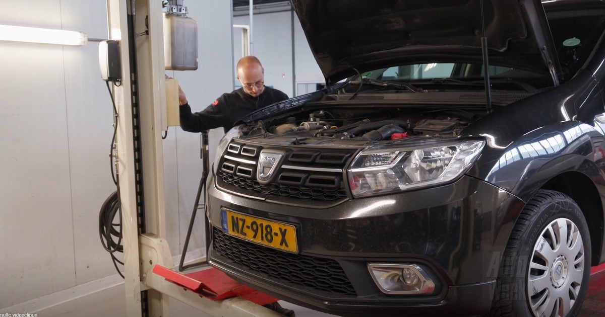 Un autoturism Dacia de culoare închisă, cu capota ridicată, pe un elevator auto într-un atelier de reparații, în timp ce un mecanic lucrează sau inspectează zona motorului.