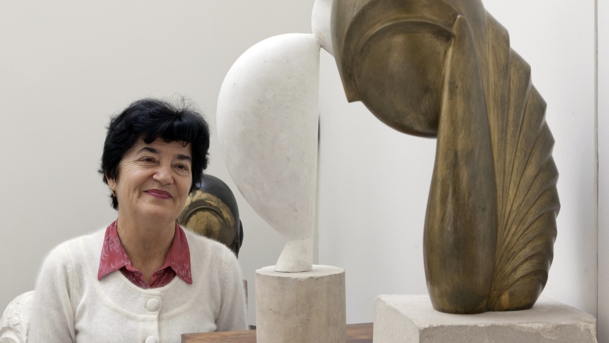 O doamna bruneta imbracata intr-un pulovar ab=lb si o camasa visinie, alaturi de cateva sculpturi ale lui Constantin Brancusi
