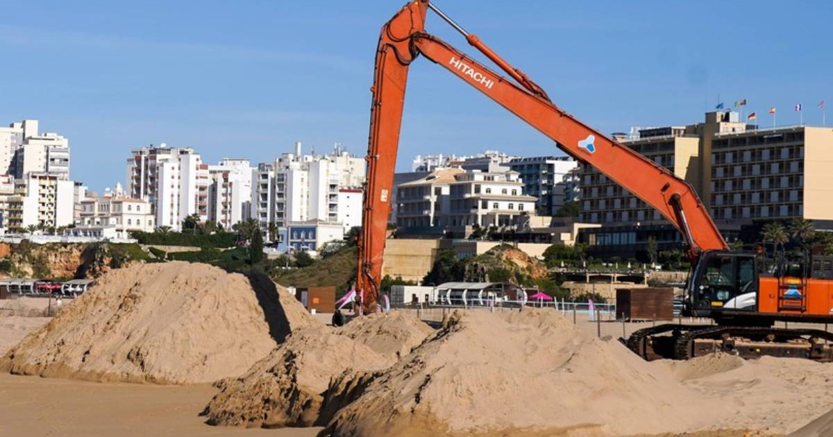 Un excavator uriaș mută nisip pe o plajă din Algarve, în fața hotelurilor și blocurilor de pe litoralul portughez.
