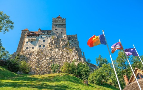 Castelul Bran, aflat pe un deal stâncos, fortăreață medievală și monument național, cu steagurile românești, ale județului Brașov și ale Uniunii Europene fluturând în față, sub un cer albastru senin în România, Transilvania