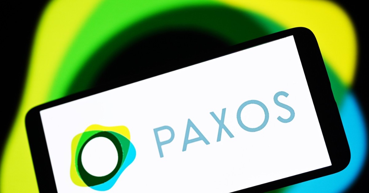 Imaginea arată sigla companiei de infrastructură blockchain Paxos pe ecranul unui smartphone, pe un fundal estompat și colorat. Sigla combină un simbol grafic modern, format din forme suprapuse de culoare galben, verde și albastru, cu numele "PAXOS" scris cu litere simple și subțiri de culoare albastru deschis.