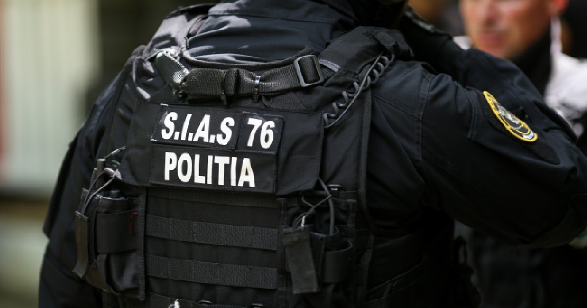 Imaginea prezintă spatele unui ofițer de poliție din forțele speciale, echipat cu o vestă tactică neagră pe care sunt vizibile inscripțiile „S.I.A.S 76” și „POLIȚIA”.