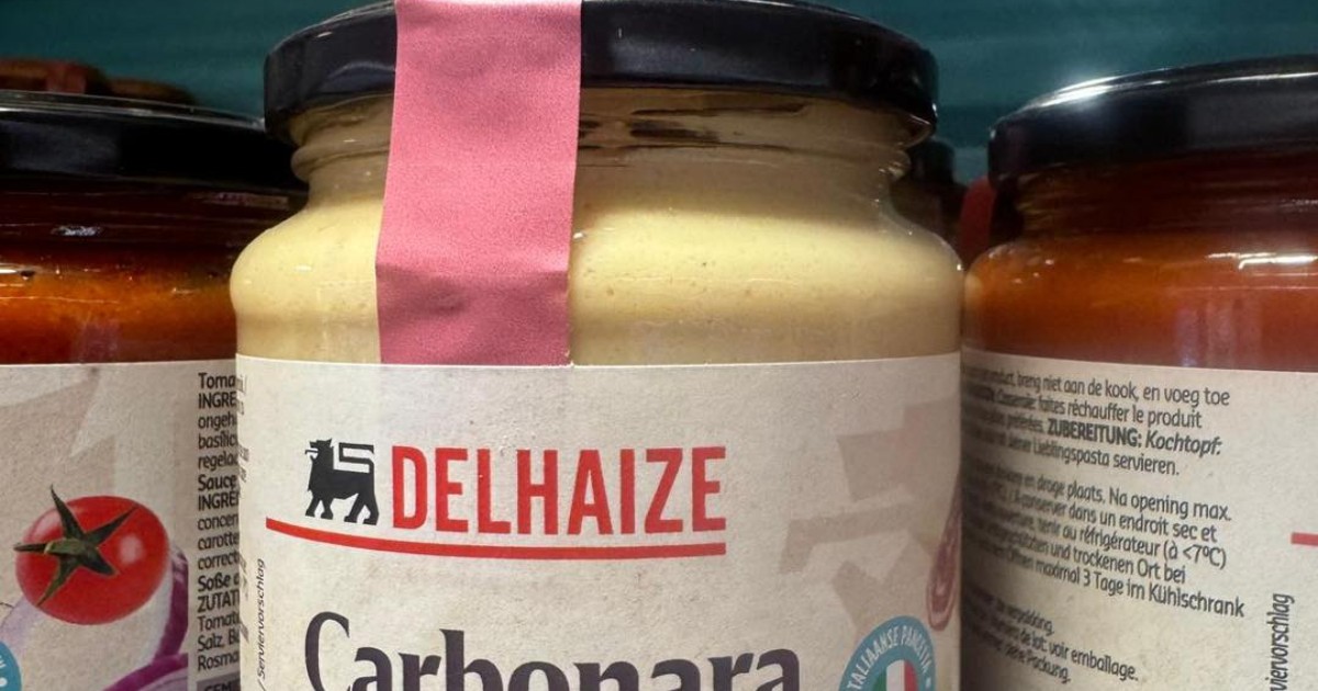 Imaginea prezintă un prim-plan al unui borcan de sticlă ce conține sos cremos Carbonara, produs de brandul DELHAIZE, eticheta fiind parțial acoperită de o bandă de sigilare roz. Sosul are o culoare galben pal, iar sub numele "Carbonara" este menționat în limba germană și franceză că este un "Sos cremos Carbonara", sugerând utilizarea în prepararea pastelor