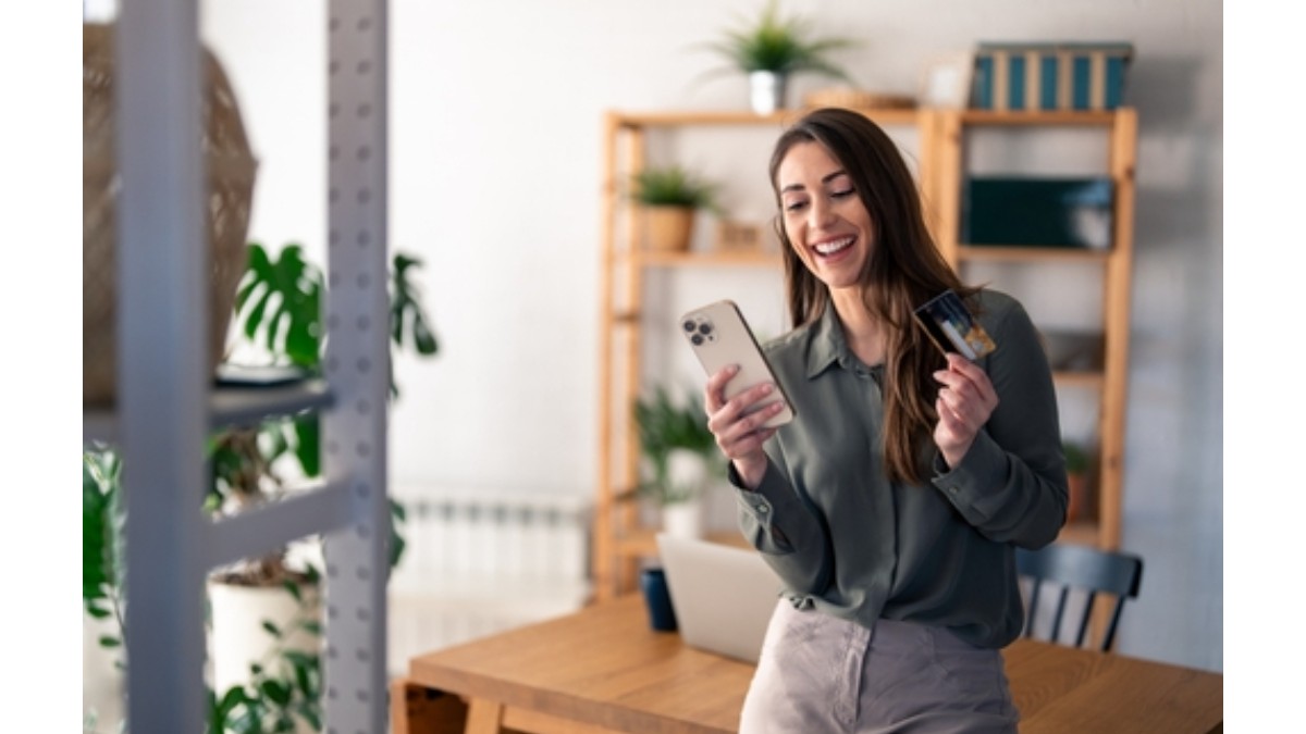 Femeie tana, fericita, imbrcata intr-o pereche de blugi gri si camasa verde smarald, tine intr-o mana un card de credit, iar in cealalta telefonul