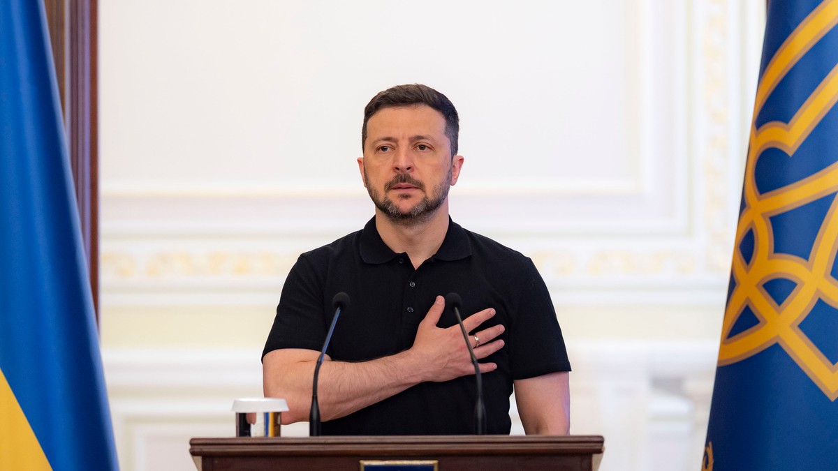 Voldimir Zelenski, îmbrăcat într-un tricou polo cu mânecă scurtă, cu mâna stângă la piept, stând la un pupitru de lemn închis la culoare pe care sunt amplasate două microfoane