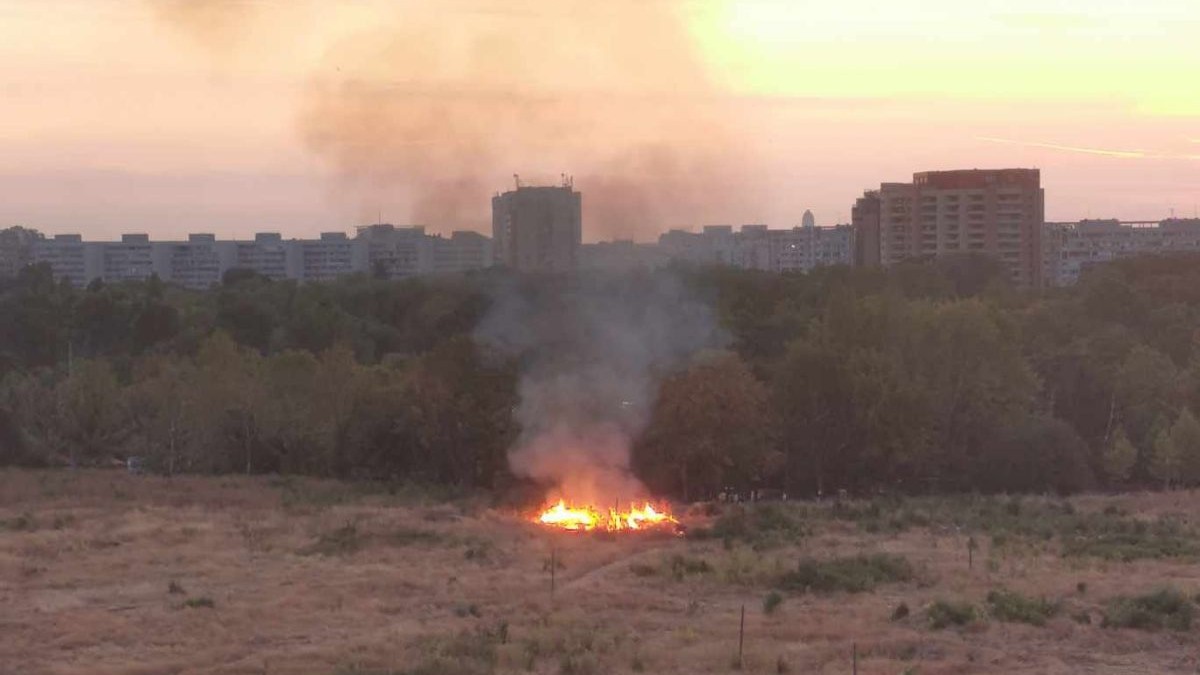 incendiu într-o zonă de vegetație uscată din zona retrocedată a parcului IOR din București. Un cerc de iarbă arde cu flăcări mari, vizibile, și degază fum gros și negru. pe fundal se vede peisajul urban al Bucureștiului, cu blocuri înalte iar pe fundal lumina soarelui denotă faptul că este apus