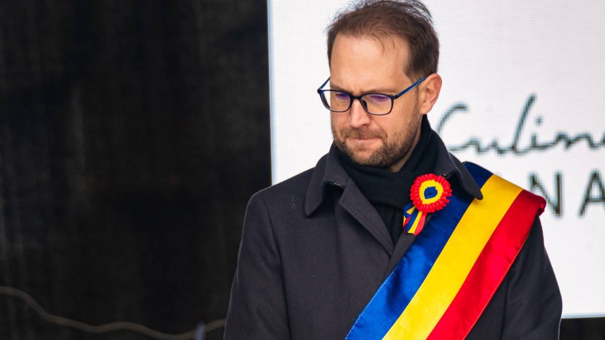 Portretul lui Dominic Fritz, primarul municipiului Timișoara, într-o haină neagră și cu steagul românesc în jurul său.