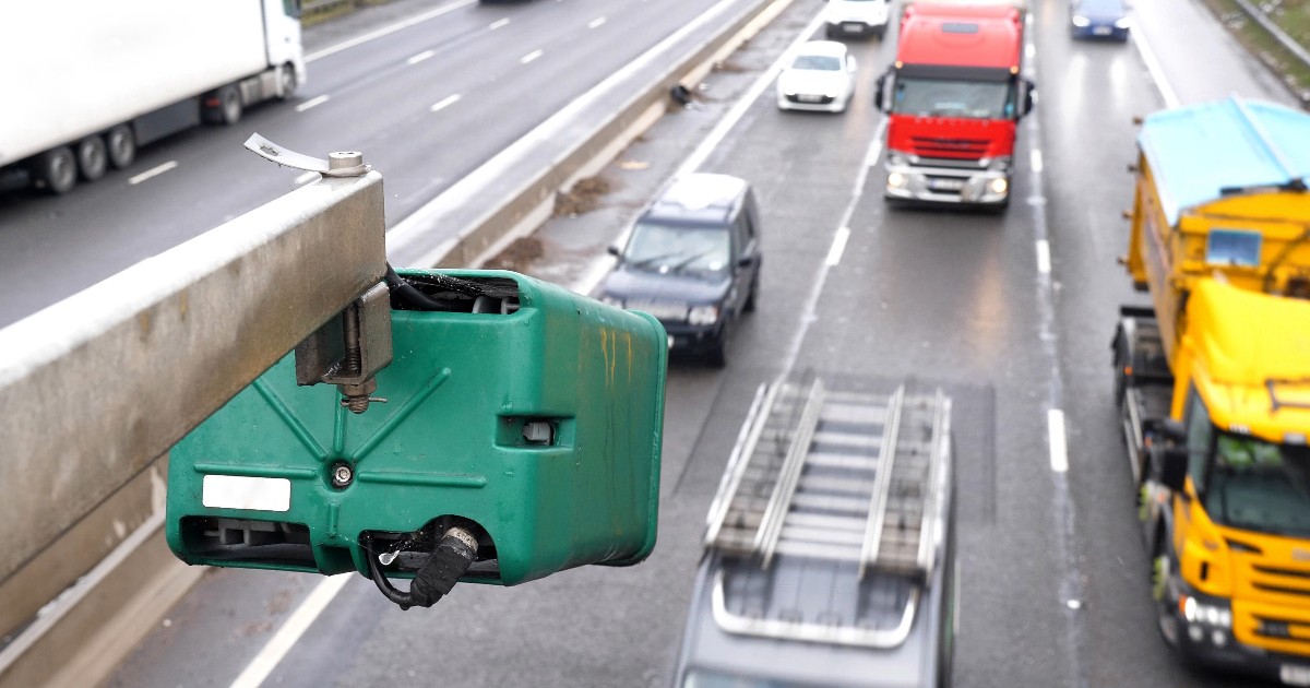 Prim-plan cu o cameră verde de supraveghere și monitorizare a vitezei montată deasupra unei autostrăzi aglomerate, pe care circula vehicule și camioane.