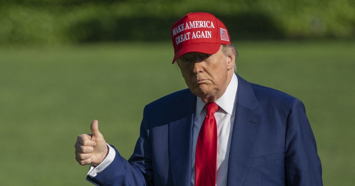 Donald Trump, îmbrăcat la costul gri, cu cravată rși și purtând pe cap o șapcă roșie cu mesajul „Make America Great Again”, ține degetul mare ridicat pe peluza din fața Casei Albe