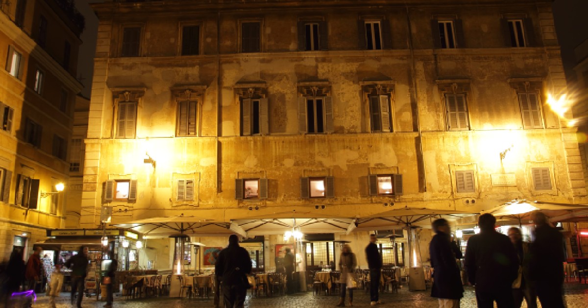 Imaginea surprinde atmosfera nocturnă a unei piețe din Roma, unde trecătorii și clienții se plimbă pe lângă terasele iluminate ale restaurantelor, aflate la parterul unei clădiri istorice cu fațadă veche.