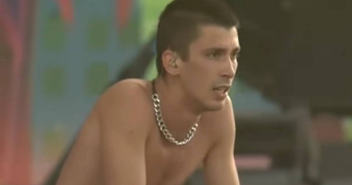 În imagine, artistul Cojo stă pe scenă privindu-și fanii. Este la bustul gol și în mână ține un microfon