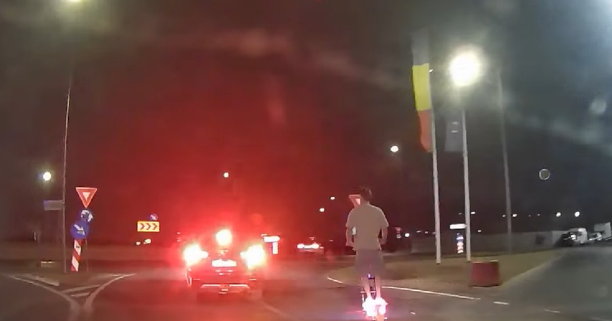 Imaginea este o fotografie de noapte, probabil realizată dintr-o mașină, și surprinde o scenă de trafic. În prim-plan se află un bărbat care merge cu o trotinetă electrică. Roata din spate a trotinetei are o lumină puternică, de culoare roz. În fața bărbatului, se află o mașină cu stopurile roșii aprinse. Pe marginea drumului, se văd semne de circulație și un stâlp de iluminat. Un steag cu culorile roșu, galben și albastru, steagul României, este vizibil în partea dreaptă.