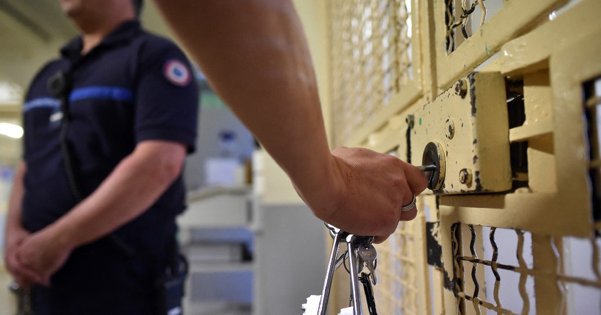 Imaginea surprinde mâna unui gardian de penitenciar în prim-plan în timp ce introduce o cheie într-o poartă metalică masivă, în timp ce un alt ofițer stă de pază în fundal.