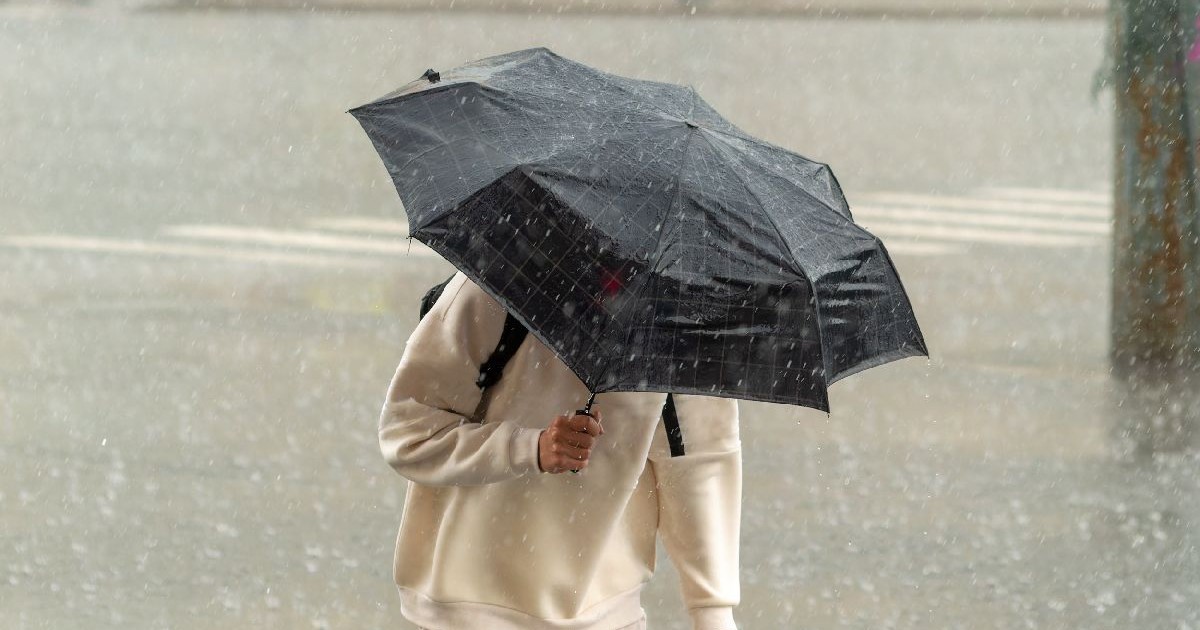 Imaginea prezintă o persoană care merge pe stradă, protejându-se sub o umbrelă neagră de o ploie torențială
