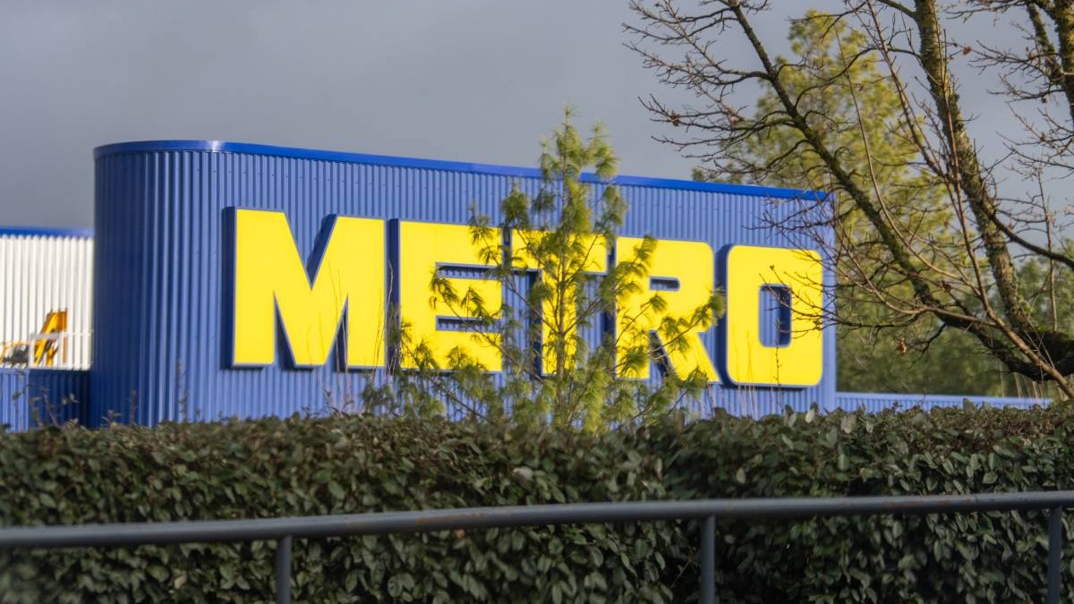 Sigla METRO, cu litere galbene strălucitoare pe clădirea albastră în spatele verdeții luxuriante