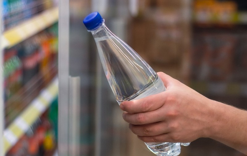 O mână care ține o sticlă din plastic cu apă și un capac albastru, scoasă sau așezată lângă rafturile frigorifice ale unui magazin.