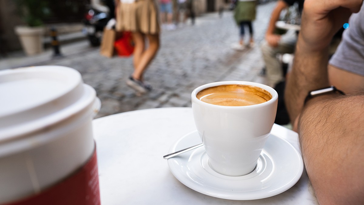 O cafea într-o ceașcă albă pe o măsuță iar în fundal se văd persoane care trec pe lângă.
