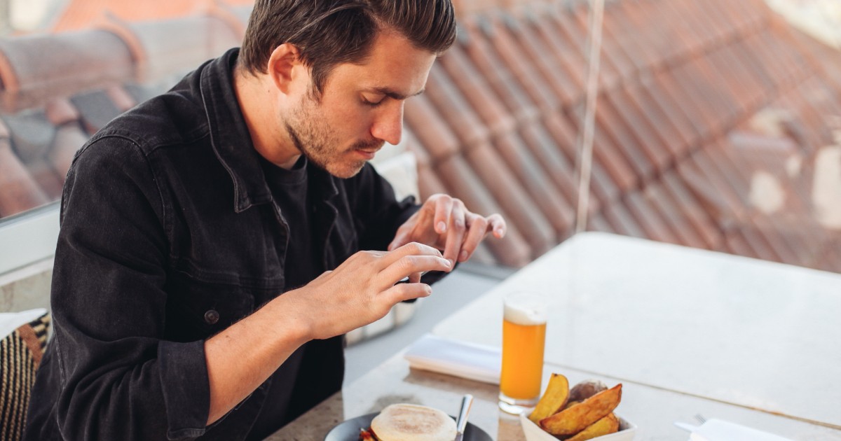 Un bărbat stă la o masă și fotografiază un burger cu telefonul mobil. În fața lui se află și o porție de cartofi și un pahar cu băutură, iar scena sugerează că verifică sau documentează preparatul înainte de a-l consuma.