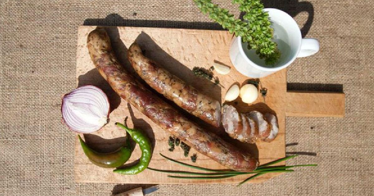 În imagine apar cârnați, ierburi aromatice, usturoi, ceapă, ardei iute și lemongrass pe o tavă rustică din lemn, cu o cană albă și un cuțit lângă ea.
