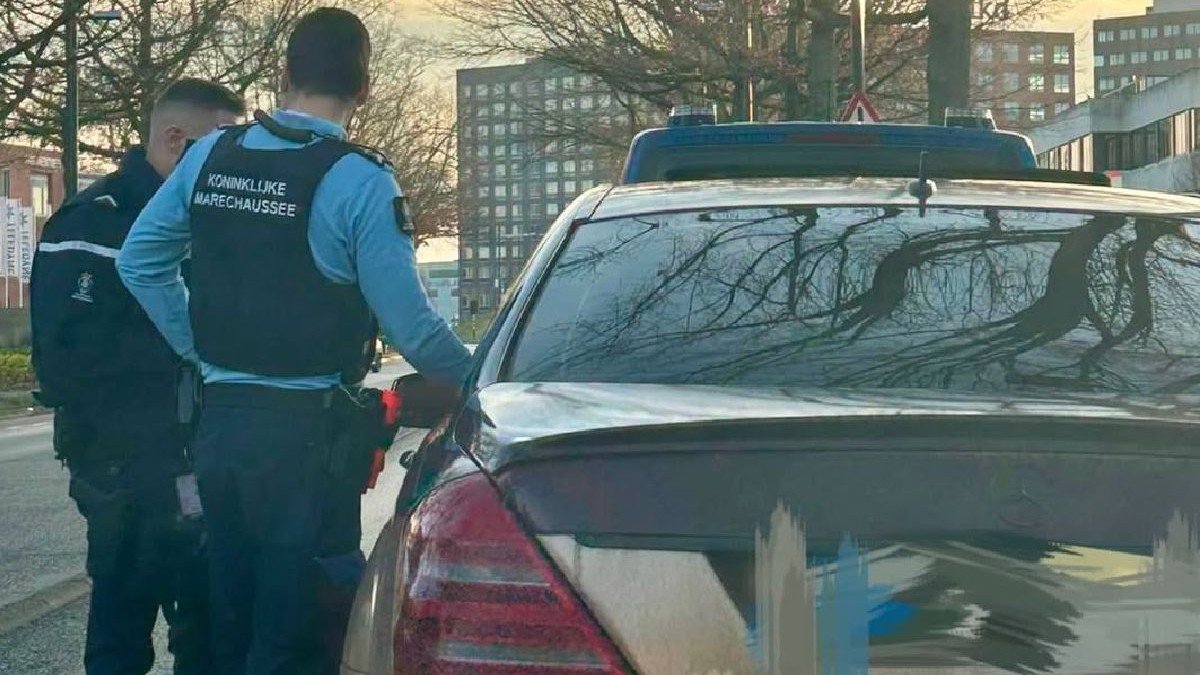 Imaginea arată doi polițiști olandezi inspectând o mașină. Autoturismul, un Mercedes-Benz, este parcat pe stradă, cu stopurile vizibile și luneta reflectând ramurile goale ale copacilor. În fundal se află clădiri.