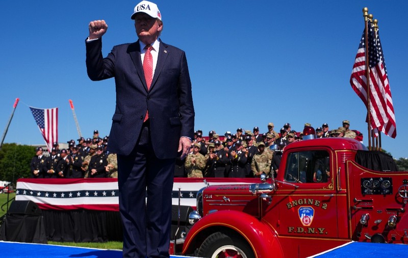 Donald Trump purtând o șapcă albă cu inscripția „USA” și ridicând pumnul, stând în fața unei mașini de pompieri roșii și a unui grup de oficiali în uniformă, pe fundalul unui cer senin.