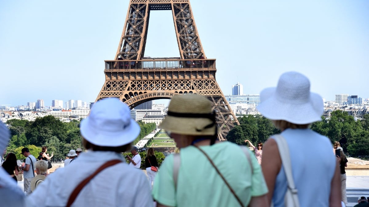 În prim-plan se află mai mulți turiști care admiră și fotografiază Turnul Eiffel, unul dintre cele mai cunoscute simboluri ale Parisului. Imaginea surprinde atmosfera unei zile însorite în capitala Franței, cu numeroși vizitatori adunați în zona de belvedere.