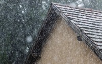 Precipitații abundente, lapoviță și ninsoare căzând peste acoperișul din țiglă al unei case, pe un fundal cu arbori.   