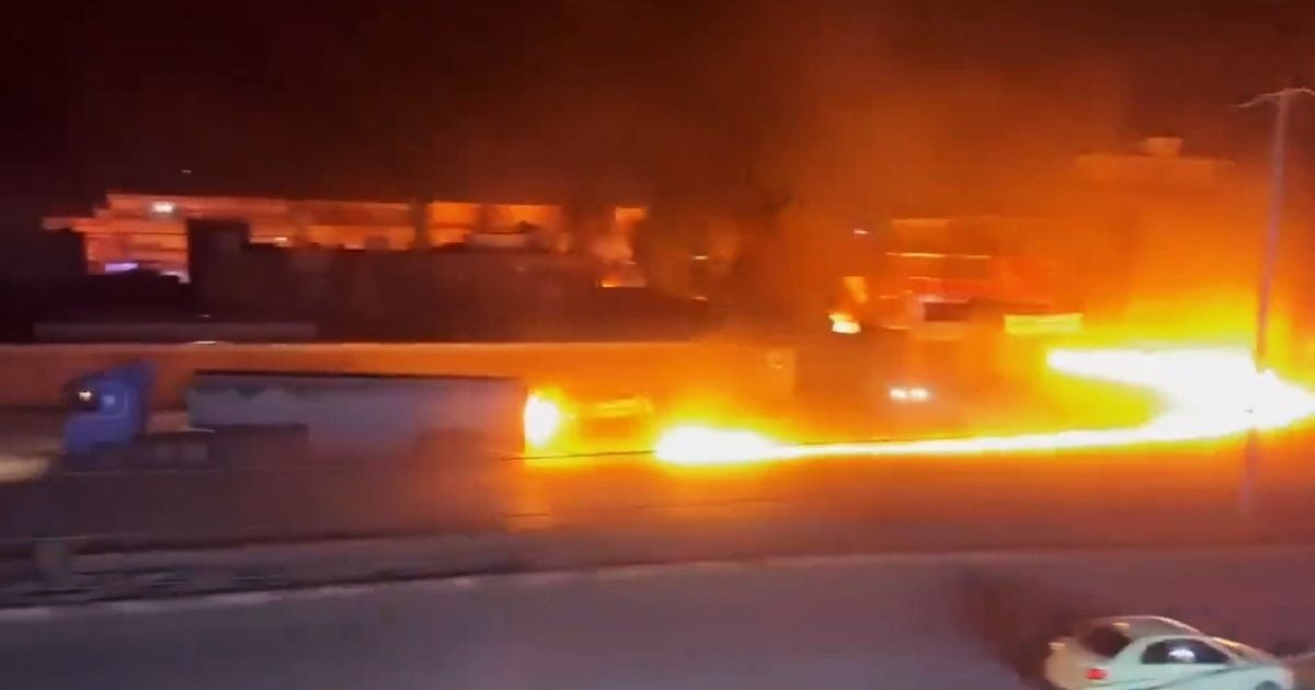 Imaginea surprinde o scenă nocturnă dramatică în care un incendiu violent se propagă sub formă de râu de foc de-a lungul unei șosele, în apropierea unui camion.