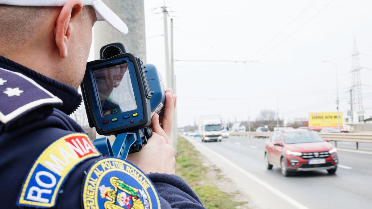 Polițist îmbrăcat în uniformă, cu șapcă albă pe cap, supraveghează traficul rutier cu un aparat radar cu laser, pe o șosea pe care circulă mașini