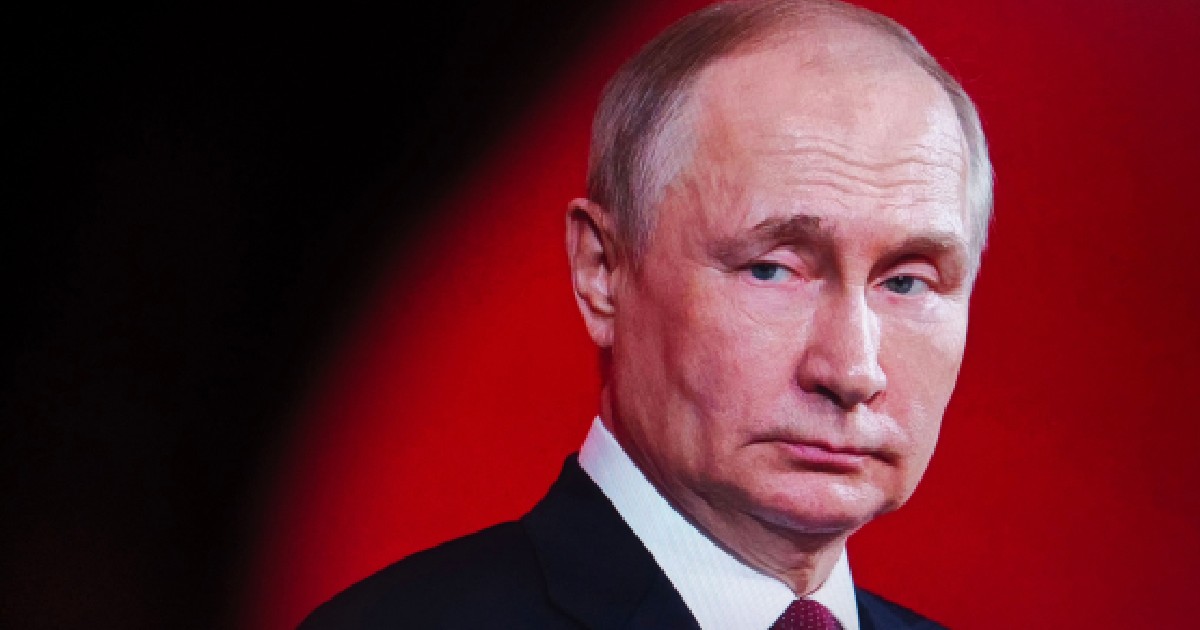 Imaginea prezintă un portret în prim-plan al lui Vladimir Putin, purtând un costum închis la culoare și o cravată vișinie, pe un fundal roșu intens.
