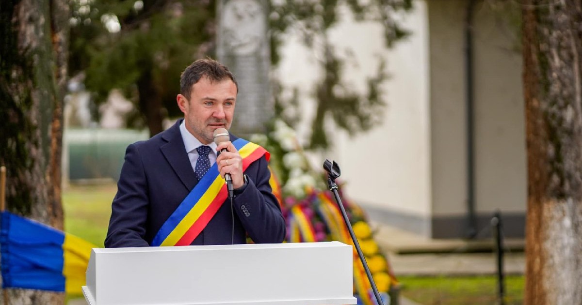 Primarul orașului Videle, Cornel Gogan, imbracat intr-un costum inchis la culoare, cămașă albă și cravată, cu panglica tricoloră peste, vorbește la microfon, în spatele unui pupitru alb, 1 decembrie 2024