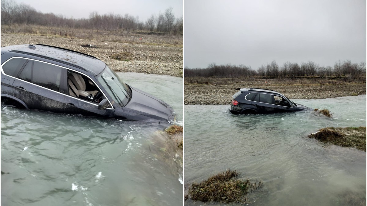 Mașina, un SUV de culoare închisă, a ajuns parțial scufundată în apă, după ce a ieșit de pe uscat și a alunecat în albia râului. Vehiculul este blocat pe jumătate în râu, în timp ce terenul din jur pare noroios și sălbatic, într-o zonă izolată.