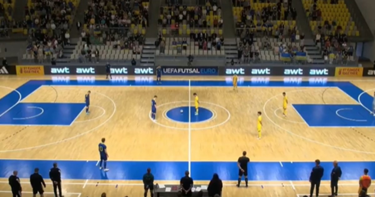 Imaginea surprinde un teren de futsal. În partea stângă sunt jucători îmbrăcați în albastru, iar în partea dreaptă sunt jucători în echipament galben. În fundal se observă tribuna cu câțiva oameni așezați pe scaune.