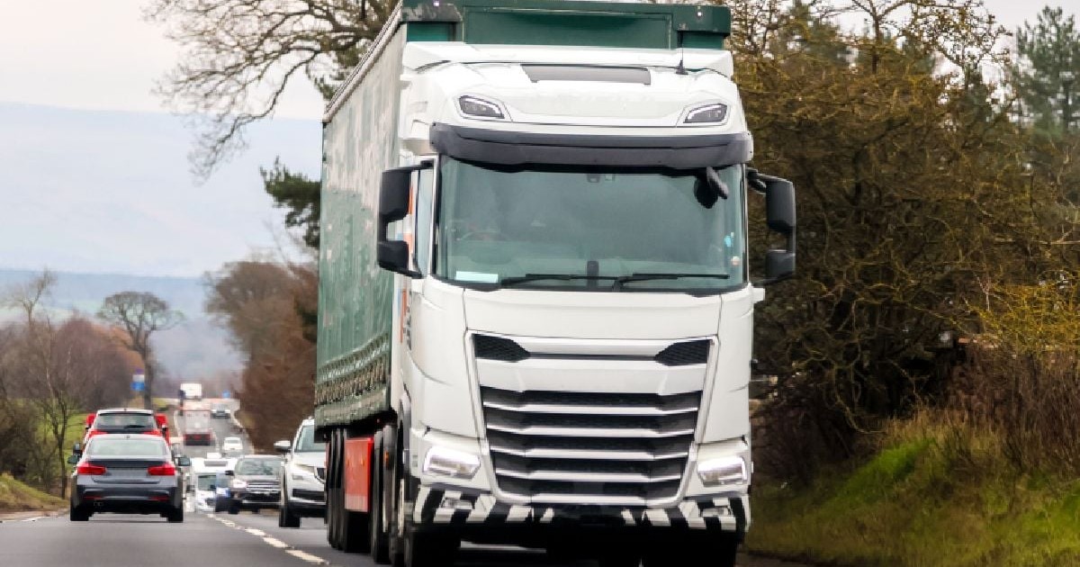 Imaginea surprinde traficul pe o șosea cu două benzi. Un camion mare, alb, care circulă spre privitor, dominând partea dreaptă a cadrului. În fundal, o coloană de mașini pe un drum șerpuit, mărginit de copaci desfrunziți și vegetație de primăvară timpurie sau toamnă.